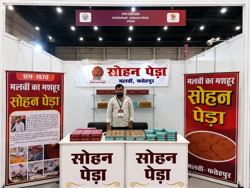Sohan Peda exhibition stall with one person behind the counter, Malwan Fatehpur banners, and boxes of peda. The overhead board reads H11-05/26, Fatehpur — Sohan Peda.
